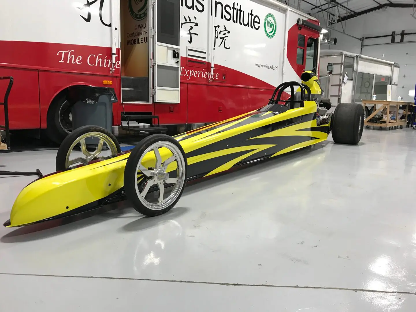 A yellow and black dragster sitting in front of a red truck.